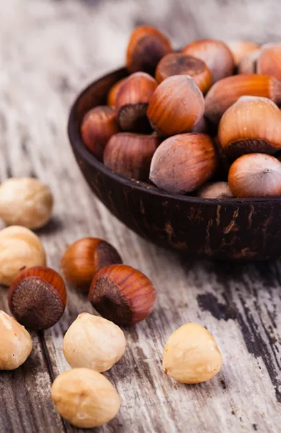 Avelãs inteiras com casca numa taça de madeira escura, com frutos descascados e não descascados sobre madeira rústica