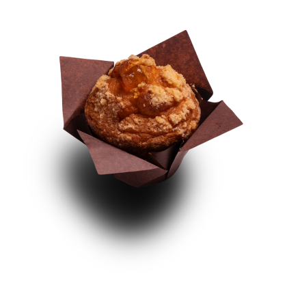 Muffin dourado coberto com crumble e recheio de fruta, envolvido em papel castanho, sobre fundo branco.